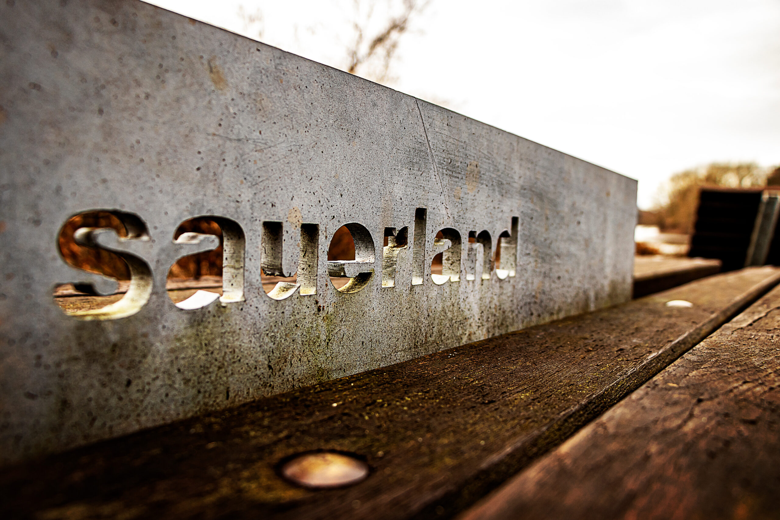 Metalltafel mit dem ausgeschnittenen Schriftzug „Sauerland“ auf einer hölzernen Sitzbank im Möhnetal, Natur im Hintergrund
