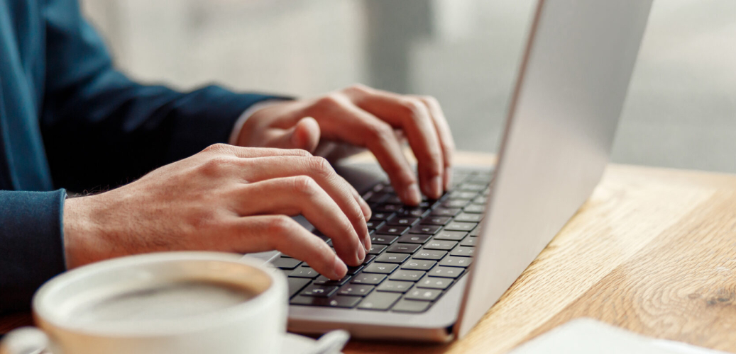 Hände tippen auf einem Laptop an einem Holztisch, daneben steht eine Kaffeetasse